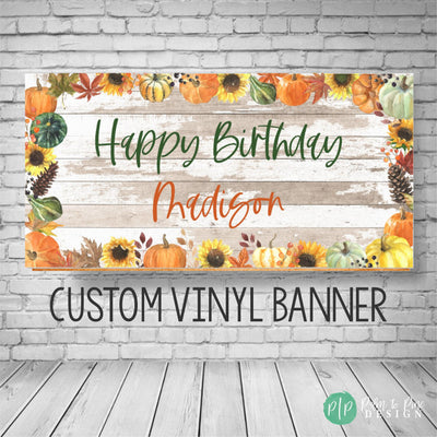 A custom vinyl banner with a fall theme. The banner is white with a woodgrain texture and features a border of orange pumpkins, green gourds, sunflowers, and fall leaves. The text on the banner reads &#39;Happy Birthday Madison&#39; in green and orange text. The banner also includes the text &#39;CUSTOM VINYL BANNER&#39;.