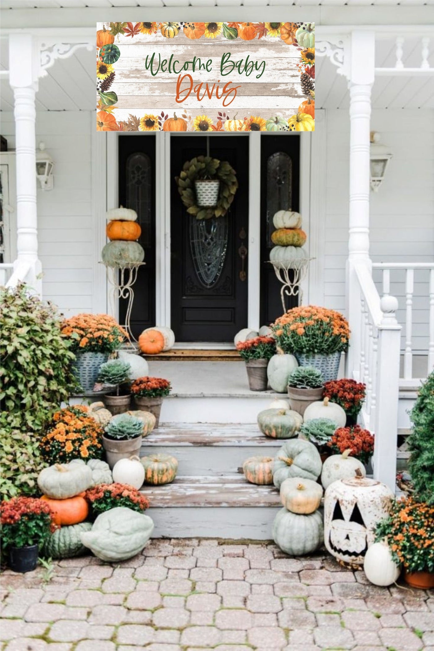 A white house with a wooden front porch decorated with a welcome baby banner with fall foliage and pumpkins. The banner reads &#39;Welcome Baby Davis&#39;. The porch is decorated with pumpkins and fall flowers in pots.