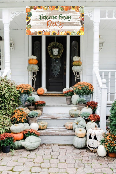 A white house with a wooden front porch decorated with a welcome baby banner with fall foliage and pumpkins. The banner reads &#39;Welcome Baby Davis&#39;. The porch is decorated with pumpkins and fall flowers in pots.