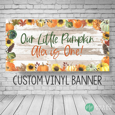A custom vinyl banner with a rustic wood background and fall-themed decorations. The banner reads &#39;Our Little Pumpkin Alex is One!&#39; with sunflowers, pumpkins, and fall leaves. The text &#39;CUSTOM VINYL BANNER&#39; is printed below the design.