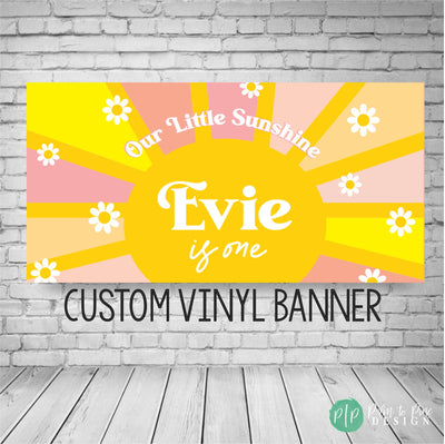 Our Little Sunshine birthday banner in yellow, pink, and tan with a sun and rays, personalized with the birthday girl’s name, and daisies scattered throughout the rays, displayed against a plain wall